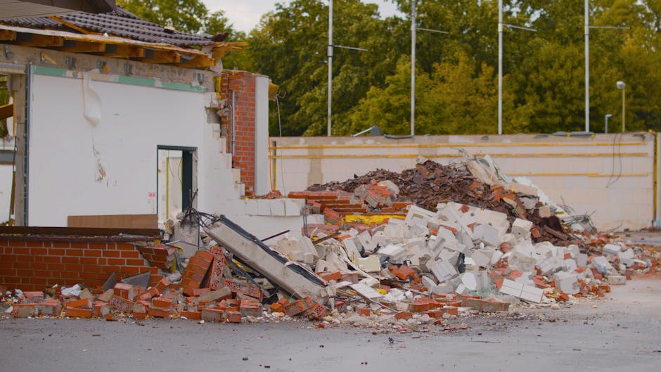 The image shows a large pile of demolition rubble situated on a paved surface in front of a partially dismantled brick and concrete structure. The rubble includes various materials such as red clay br