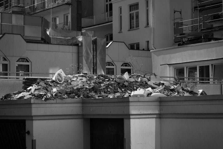 The image shows a large pile of demolition rubble situated on a paved surface in front of a partially dismantled brick and concrete structure. The rubble includes various materials such as red clay br