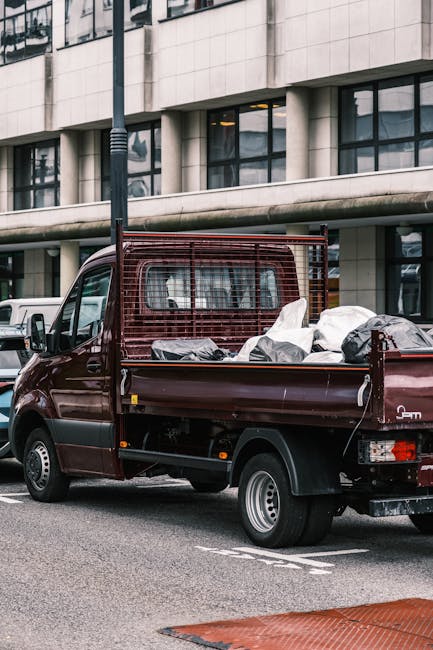 A small burgundy flatbed truck parked on an urban street in front of a modern office building with large windows framed by concrete panels. The truck's cargo bed is filled with various waste items, in